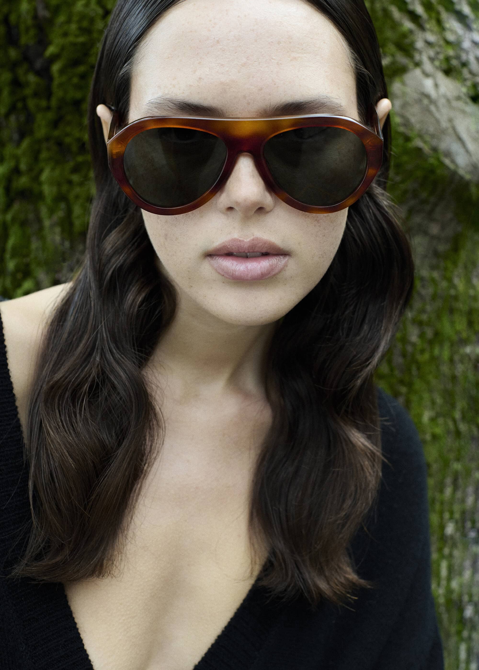 Close-up of a woman with brown hair wearing brown sunglasses, posing in front of a green leafy background in natural outdoor lighting.