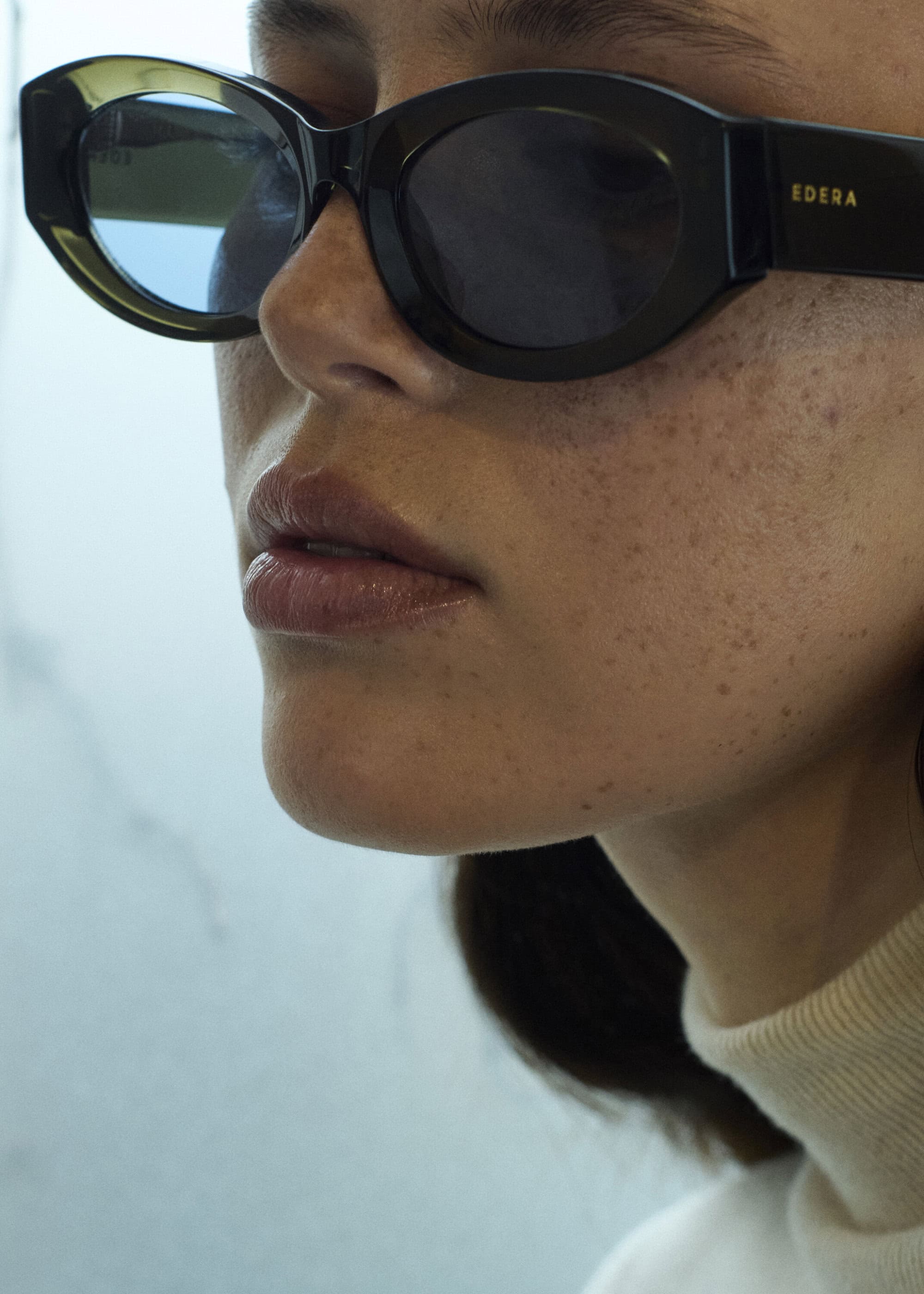 Close-up of a woman's face wearing black, oval-shaped sunglasses, featuring freckled skin and a light-colored top against a neutral backdrop.