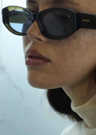 Close-up of a woman's face wearing black, oval-shaped sunglasses, featuring freckled skin and a light-colored top against a neutral backdrop.