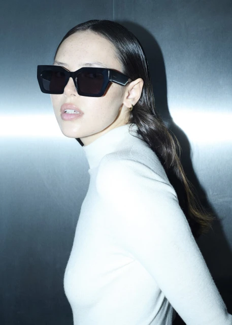 Portrait of a young adult woman wearing stylish black sunglasses and a white turtleneck against a gradient metal background.