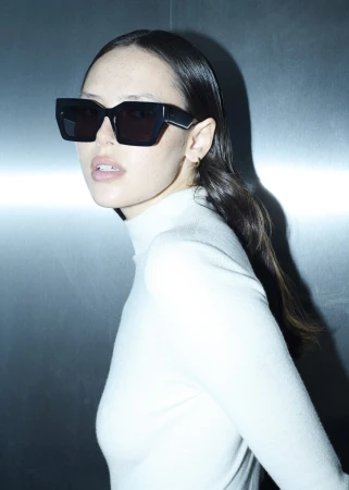 Portrait of a young adult woman wearing stylish black sunglasses and a white turtleneck against a gradient metal background.