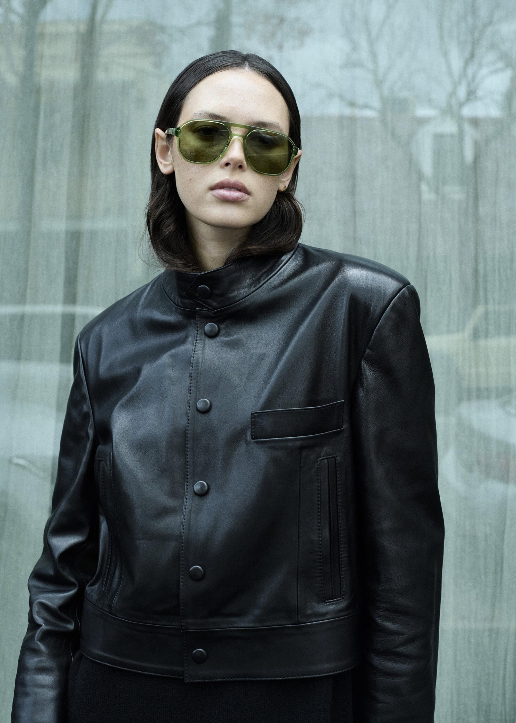 Model wearing green tinted sunglasses and a black leather jacket against an urban glass window background