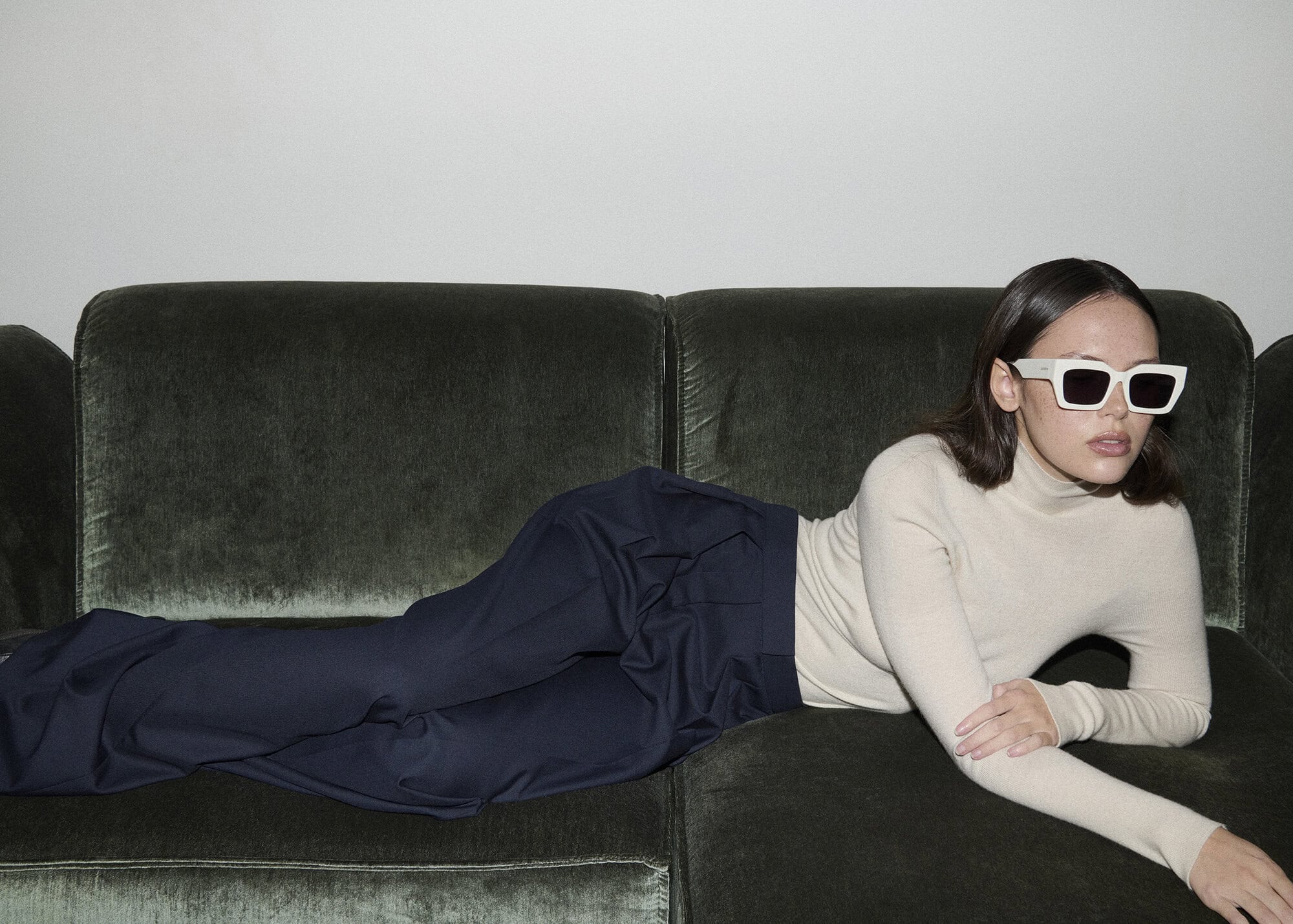 Adult female with sunglasses reclines on a dark green velvet sofa indoors, wearing a beige turtleneck and navy blue trousers.