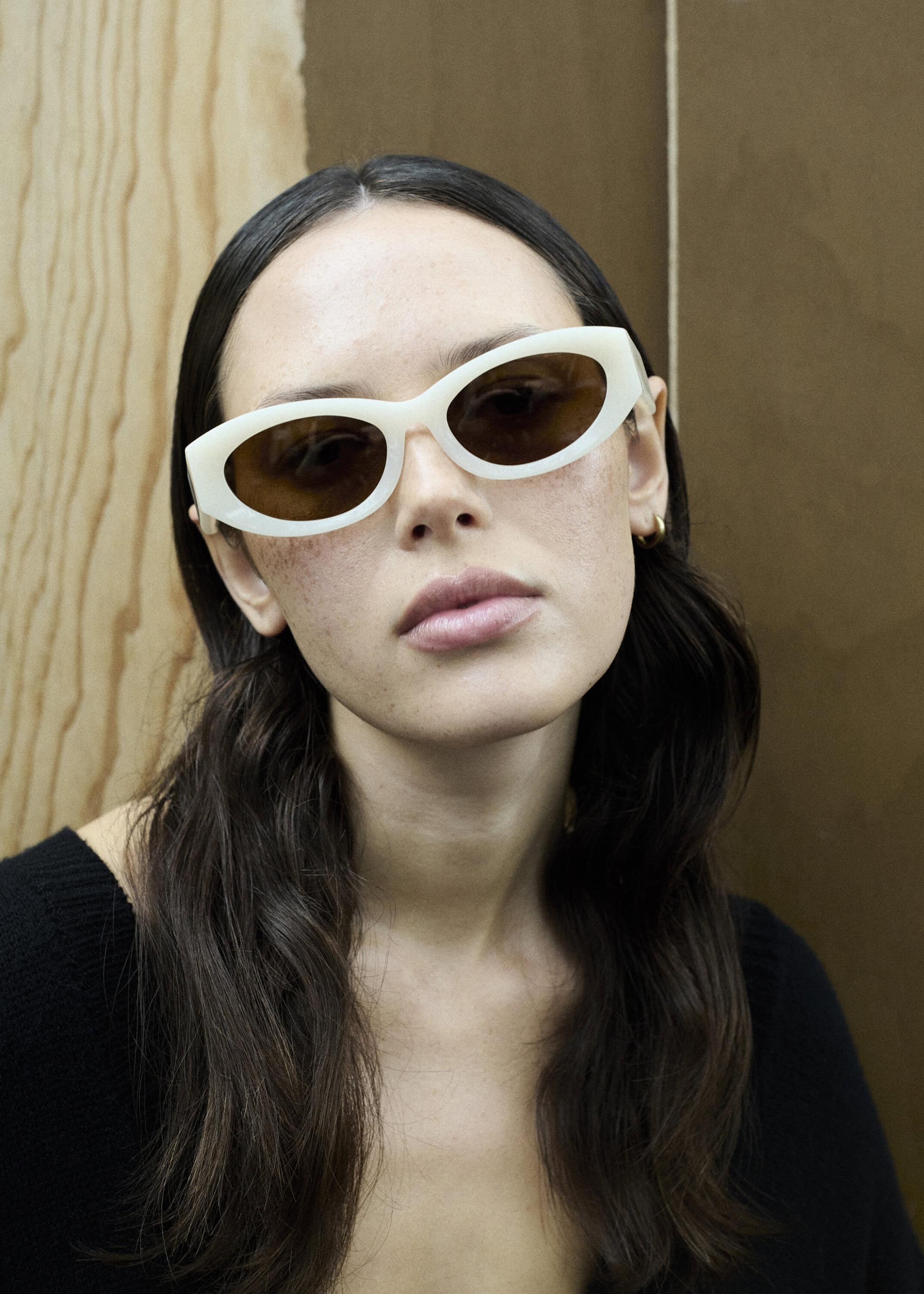 Close-up studio portrait of an adult woman with long dark hair wearing stylish cream-colored sunglasses and a black top.