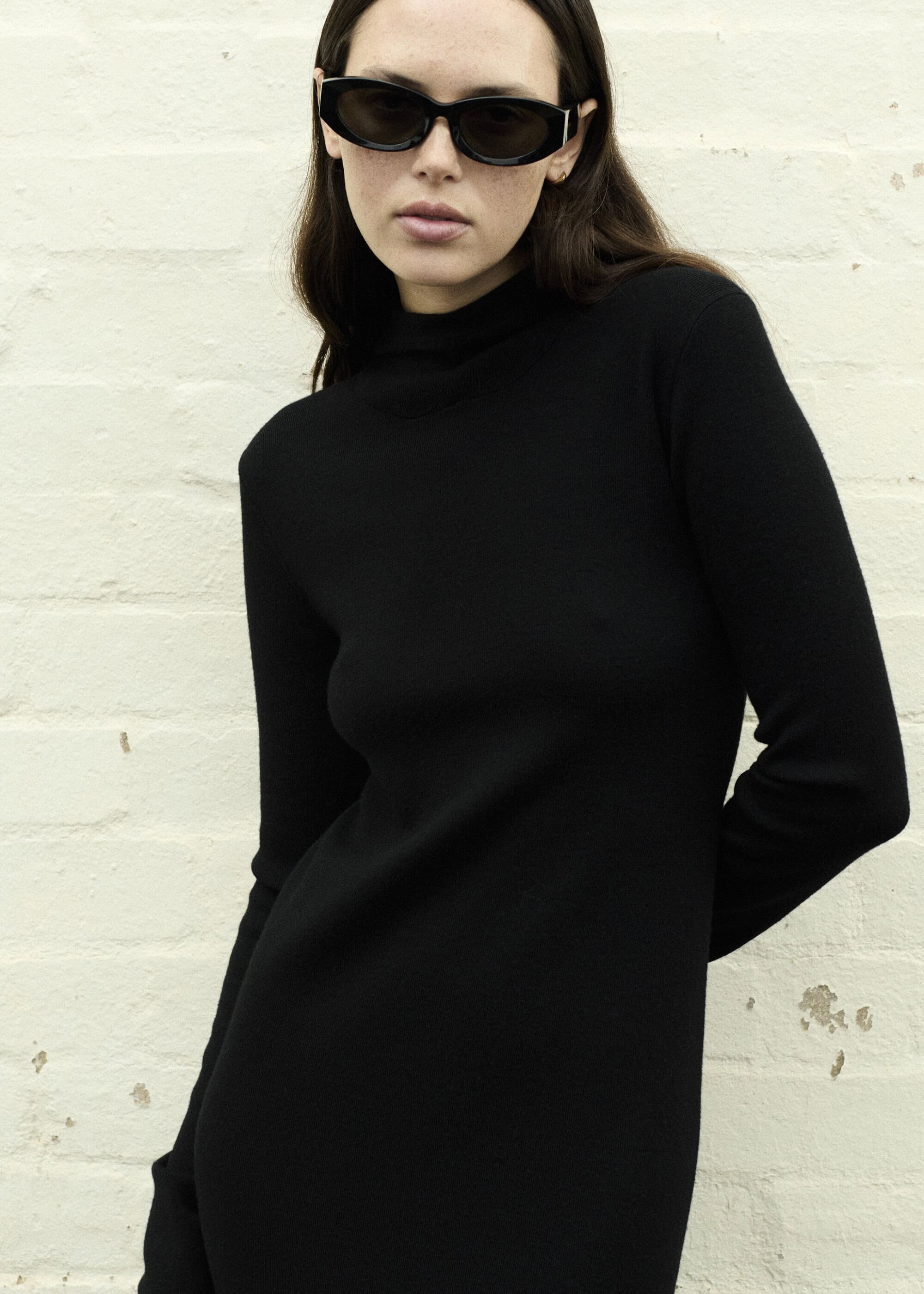 Woman in a black turtleneck dress and sunglasses stands against a white brick wall in a medium shot.