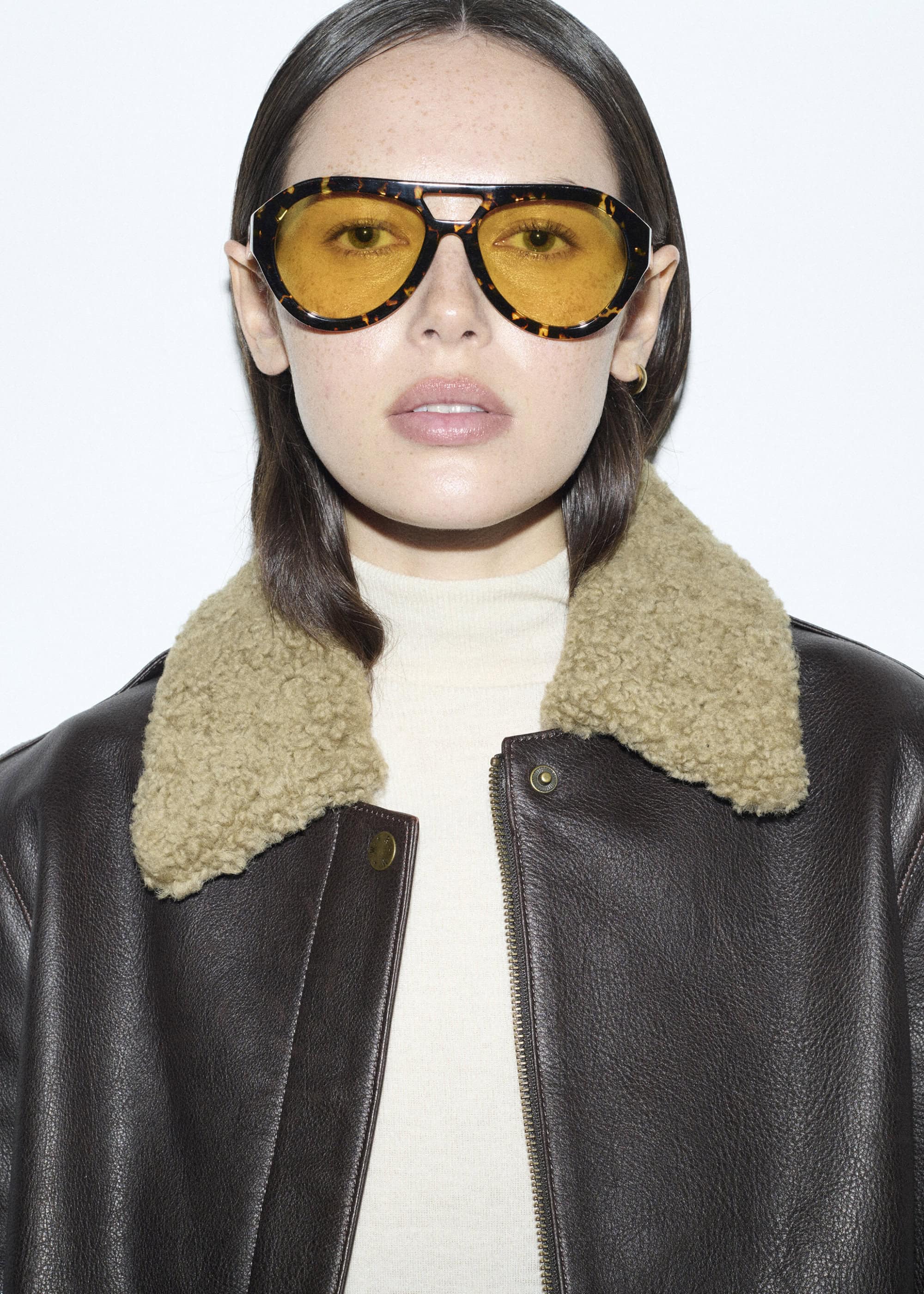Portrait of a young adult woman with brown hair wearing sunglasses and a shearling-lined leather jacket in front of a white background.