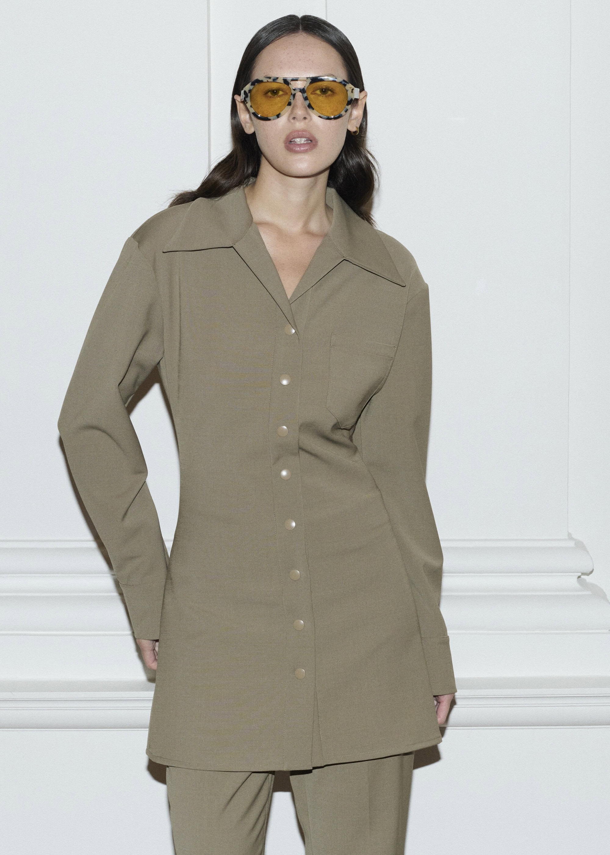 Woman in olive-green button-down shirt and matching pants poses against a white wall, wearing vintage sunglasses with yellow lenses.