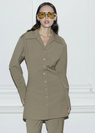 Woman in olive-green button-down shirt and matching pants poses against a white wall, wearing vintage sunglasses with yellow lenses.