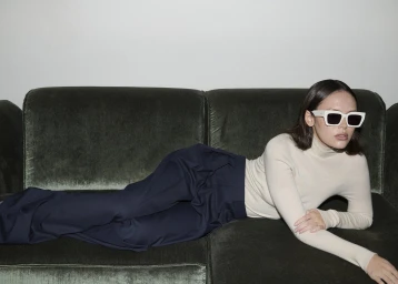 Model wearing white sunglasses and cream turtleneck lying on a dark green velvet couch