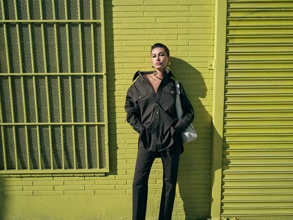 Woman in black shirt jacket and pants stands by yellow brick wall and metal gate with white tote bag over her shoulder.