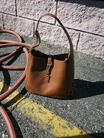 Studio shot of a brown leather handbag with gold logo hardware resting on a concrete surface outdoors with brick wall backdrop.