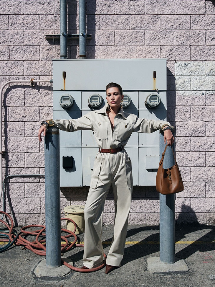 A woman in a khaki jumpsuit poses between gray metal posts, leaning on them, with an electrical box and brick wall in the background.