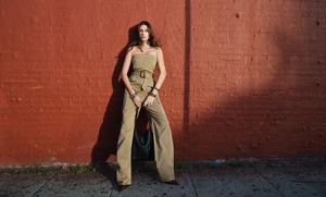 Hailey Bieber wearing a belted khaki jumpsuit leaning against a weathered red brick wall holding a black leather shoulder bag
