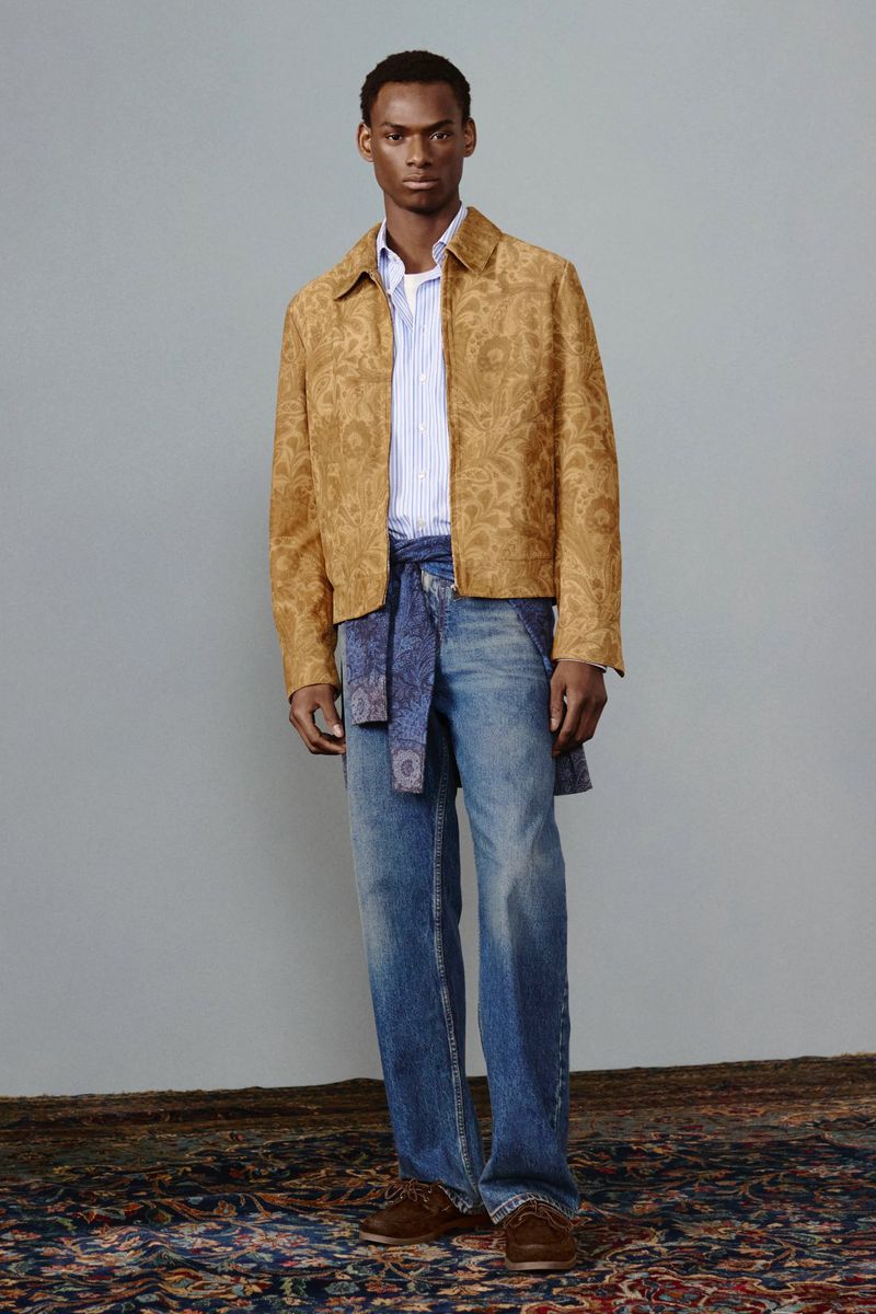 Full-length shot of an adult male wearing a patterned jacket, jeans with a blue belt, and brown shoes, posing in a studio.