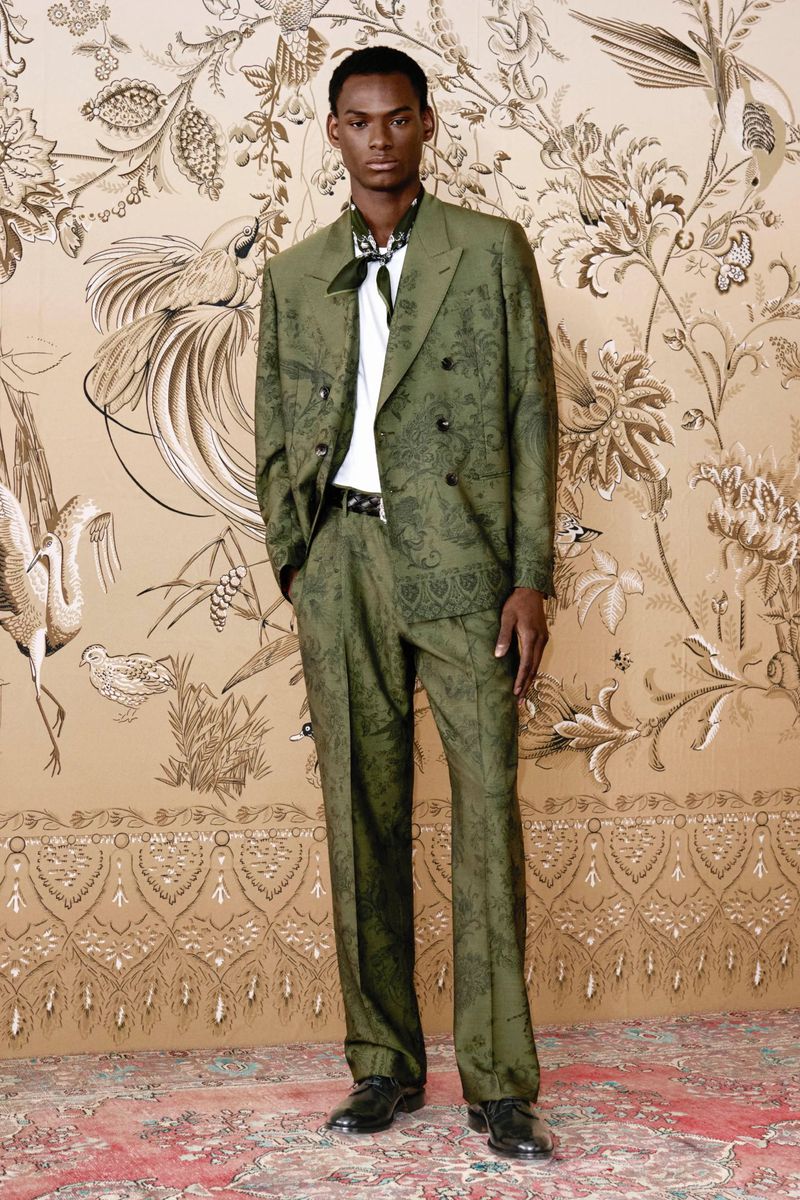 A single adult male model posing in an olive green patterned suit against a matching wallpaper with bird and floral designs.