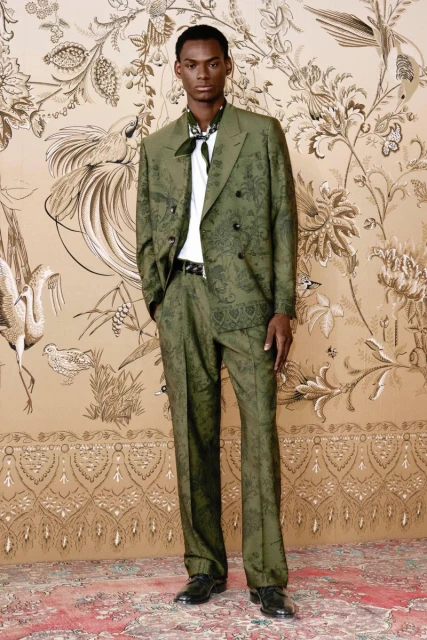 A single adult male model posing in an olive green patterned suit against a matching wallpaper with bird and floral designs.