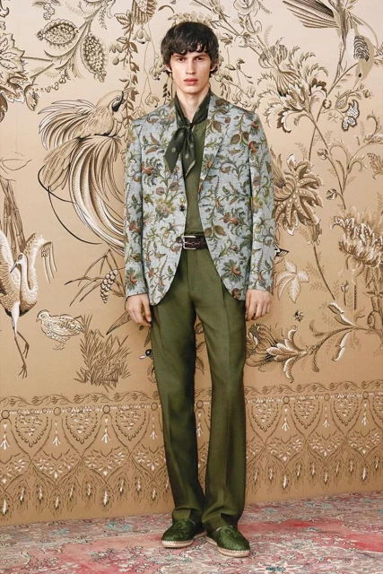 Male fashion model poses in a floral suit with a scarf against a wallpaper backdrop in a full shot, showcasing a high-fashion aesthetic.