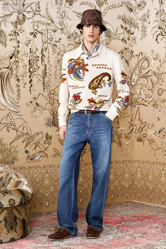 Model wearing patterned bucket hat, printed knit shirt, and denim jeans standing against illustrated wallpaper
