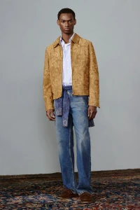 Model in patterned tan jacket and blue jeans standing against a plain grey studio backdrop over a patterned rug.