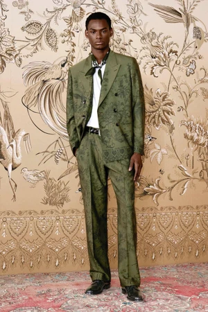 Model standing in an olive green patterned double-breasted suit against a decorative botanical wallpaper background.