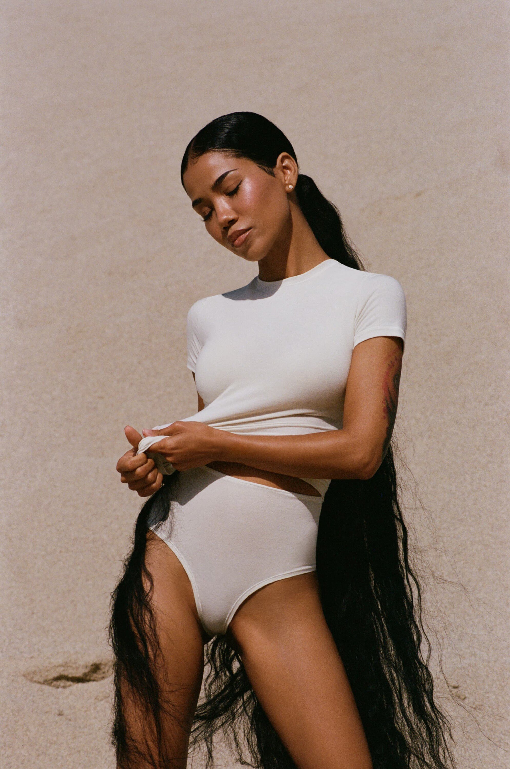 Woman with very long dark hair wearing a cream colored bodysuit against a beige sand dune background in natural light.