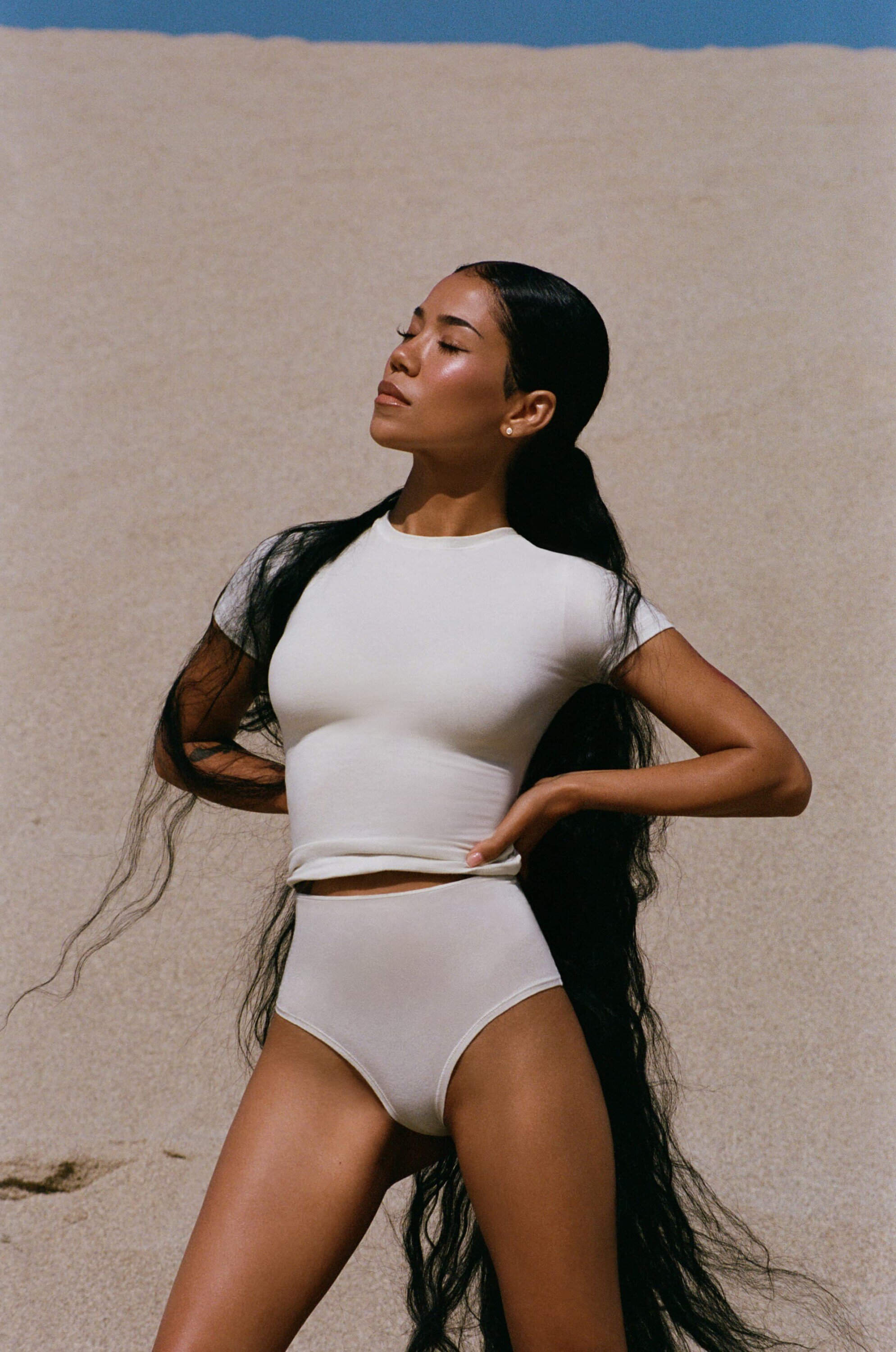 Woman with long dark hair poses in a matching ivory-colored top and briefs on a sandy beach with a clear sky in the background.
