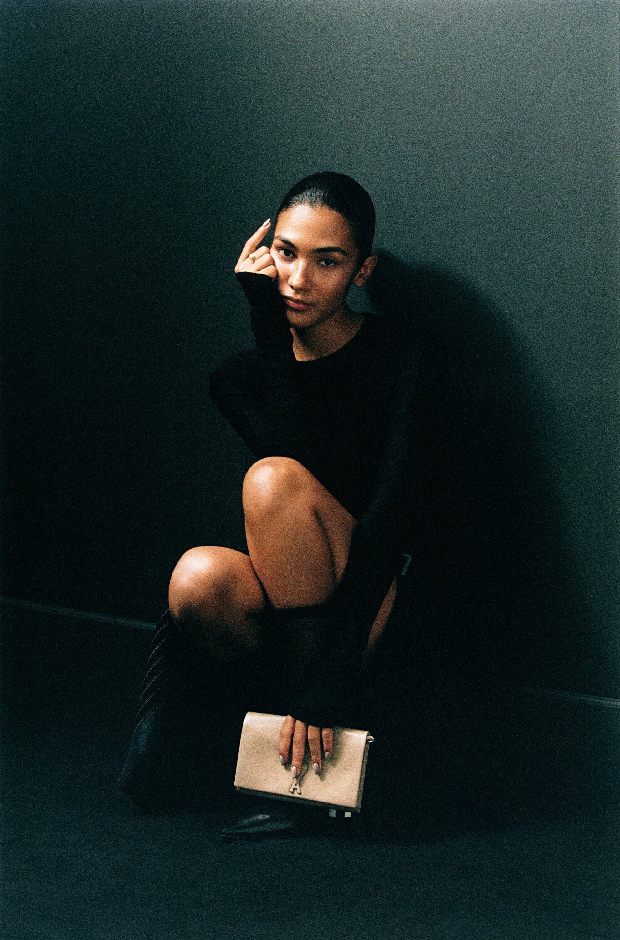 Portrait of a young woman squatting in a dark room wearing a black outfit and holding a beige clutch bag, looking at the camera.