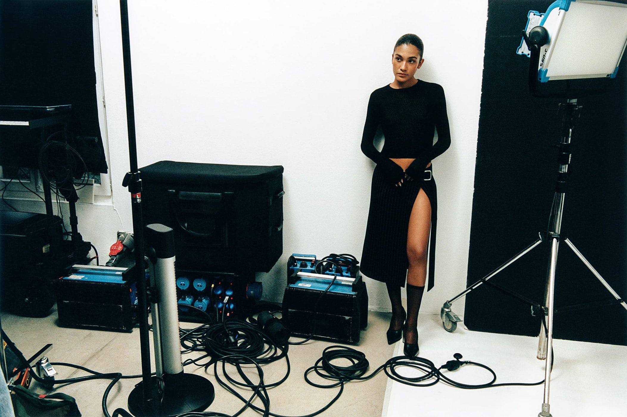Woman poses in a black dress and heels in a photo studio, alongside lighting equipment, cords, and a black and white backdrop.