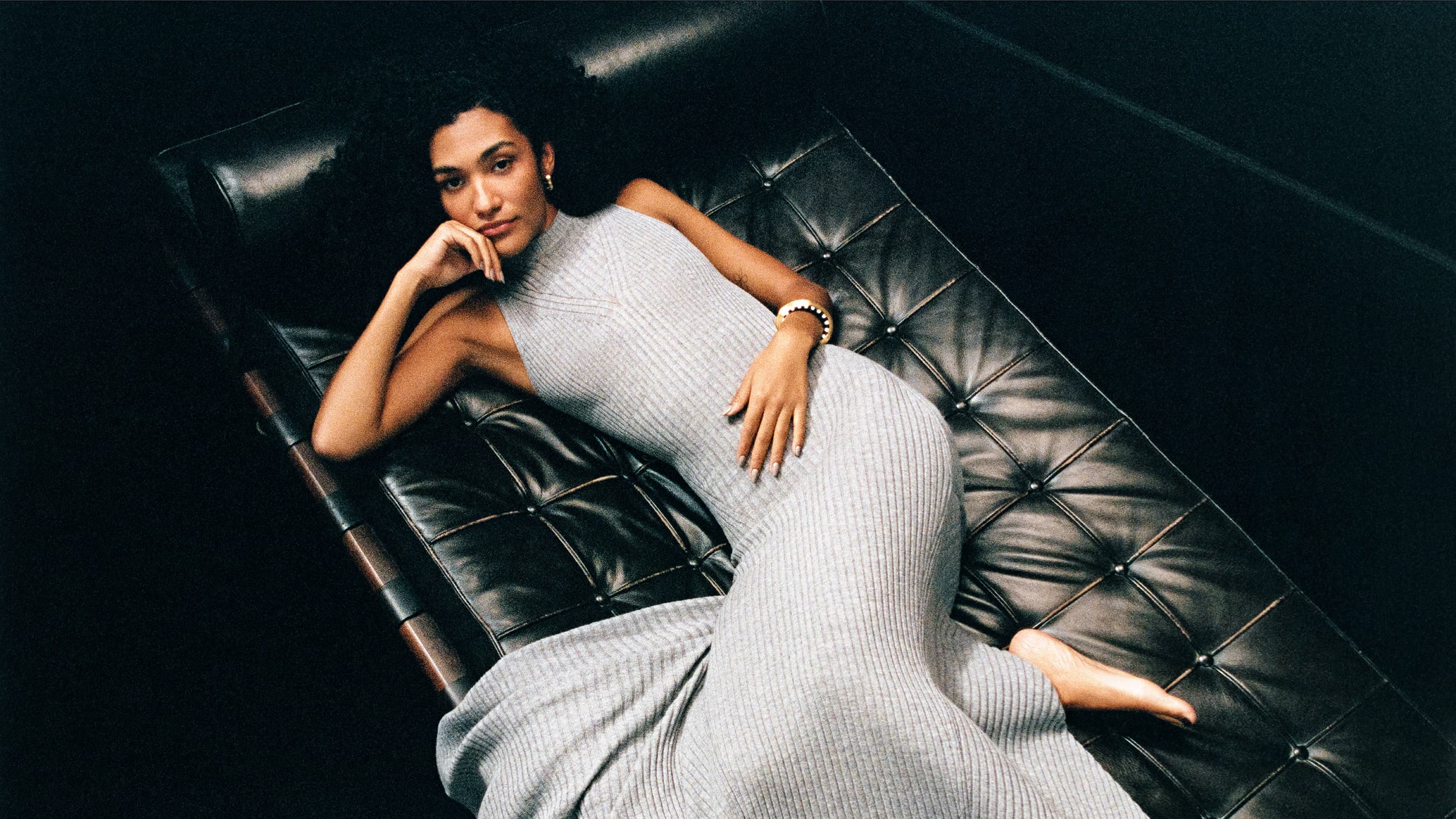 Woman in grey dress lounges on a tufted leather bench in a dimly lit indoor space, showcasing a relaxed pose and elegant fashion.