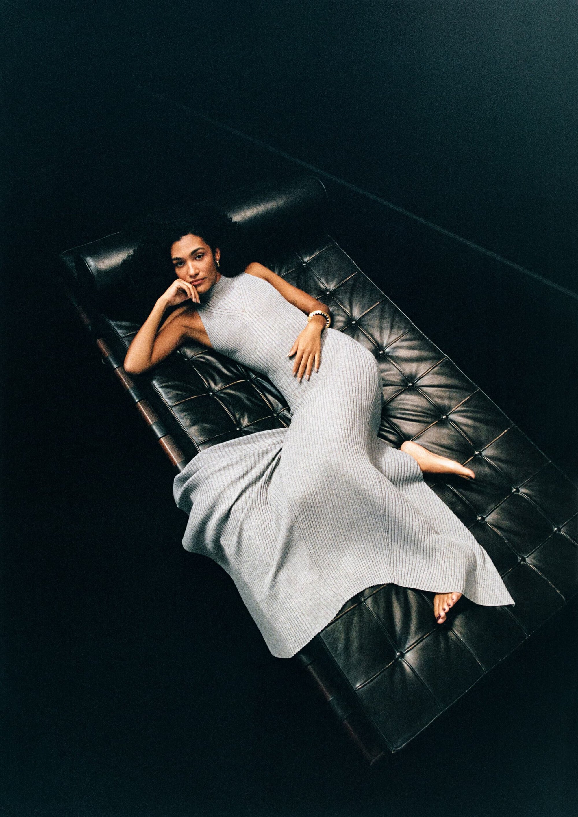 A woman with dark hair and a knit dress poses on a black leather couch in a high-angle portrait. Black interior background.