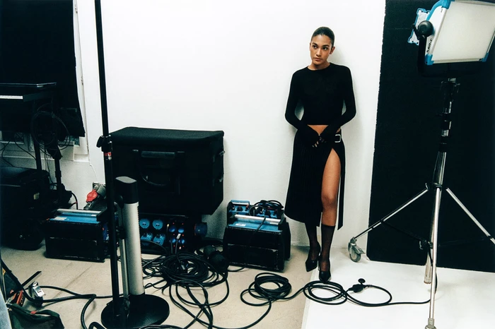 Model standing against a white studio wall surrounded by photography lighting equipment and cables for Ami.
