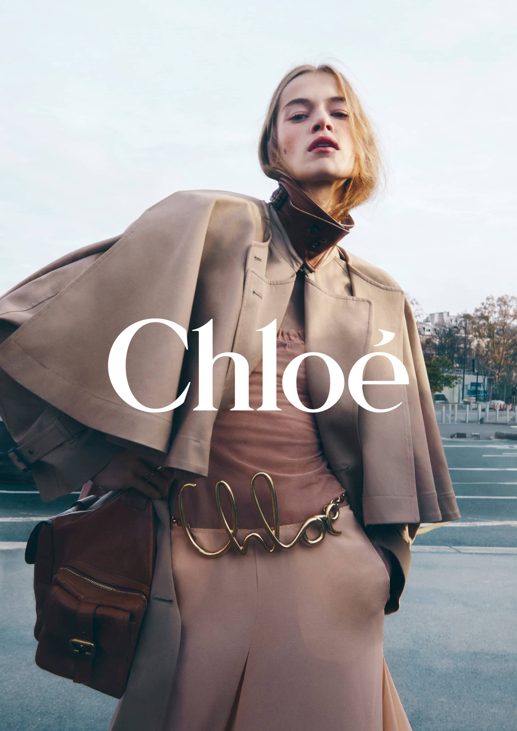 Fashion shot of a female model in a tan trench coat with a Chloe logo belt, against a blurred city background, looking at the camera.