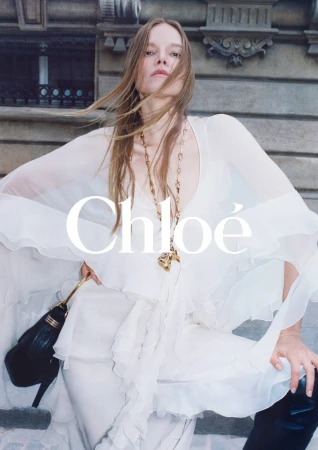 Fashion editorial shot of a blonde adult woman in a white dress and black boots, posing against an urban building with the Chloé logo.