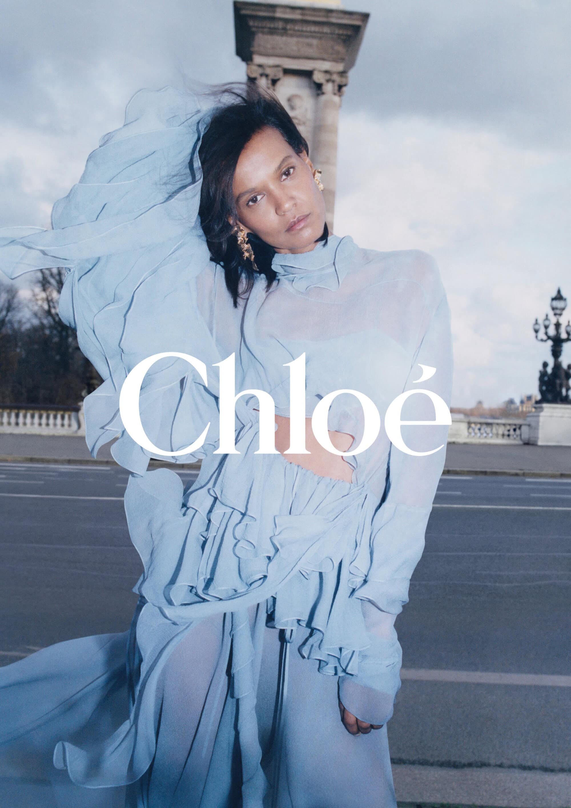 A woman in a light blue dress stands on a bridge with a column in the background. The Chloe logo is overlaid on the image.