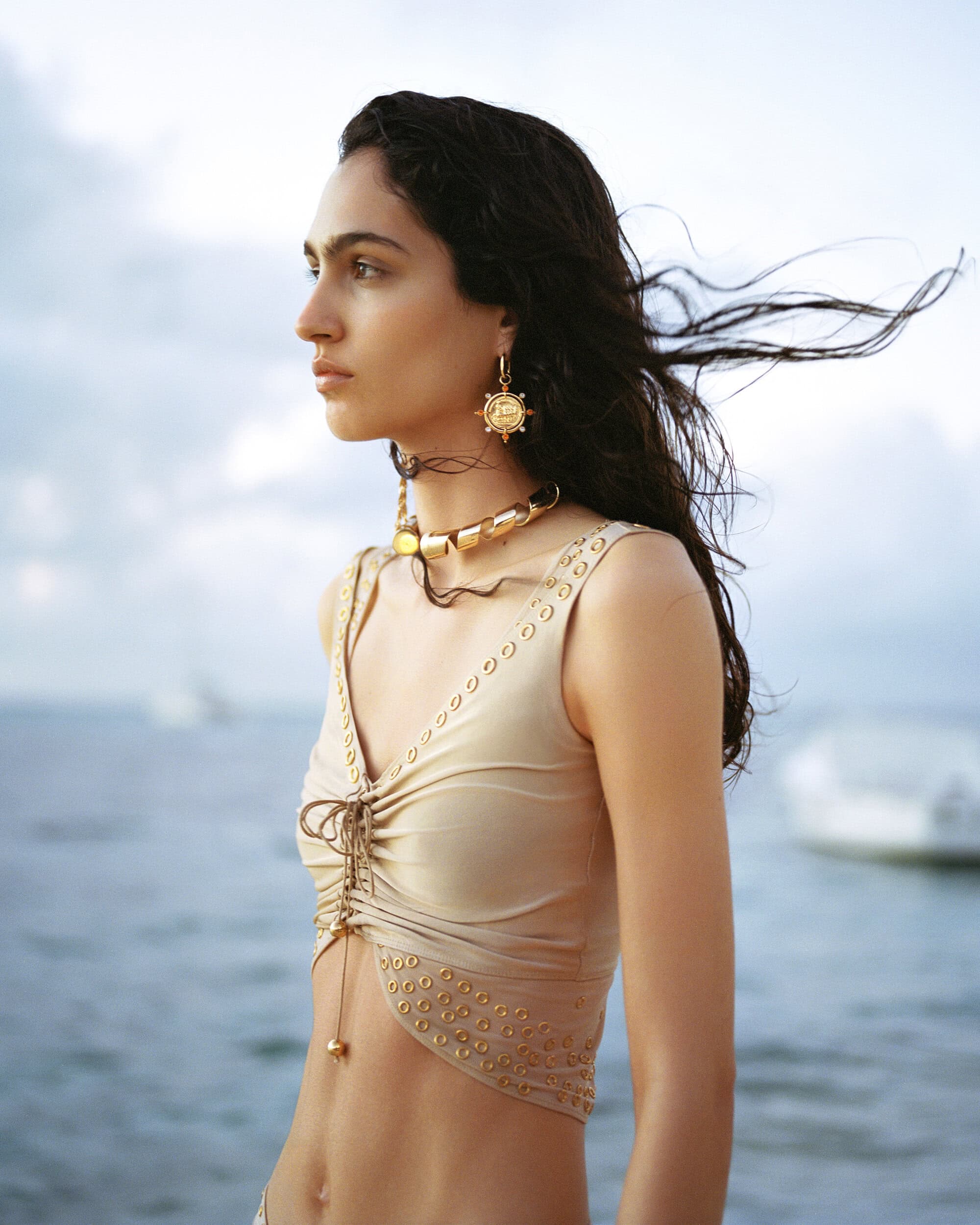 Woman in a beige two-piece and gold jewelry stands on a body of water with wind blowing her dark hair in a fashion shot.