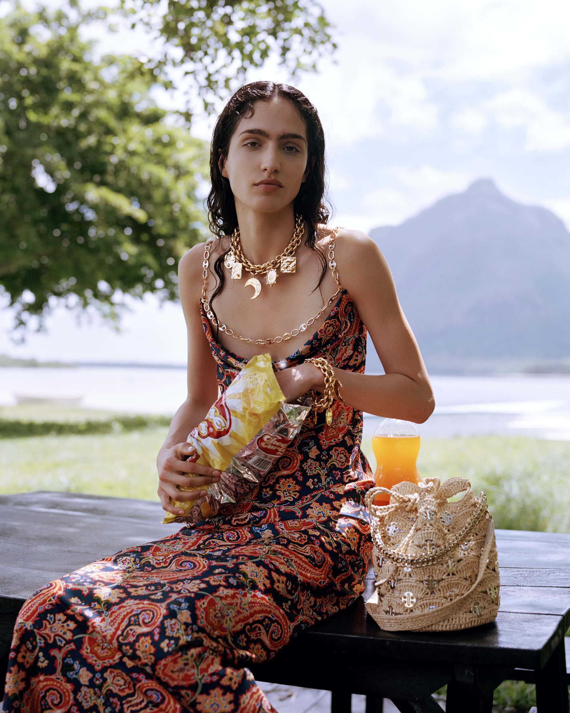 A brunette woman wearing gold jewelry and a patterned dress sits on a bench outdoors, holding a bag of chips with a lake background.