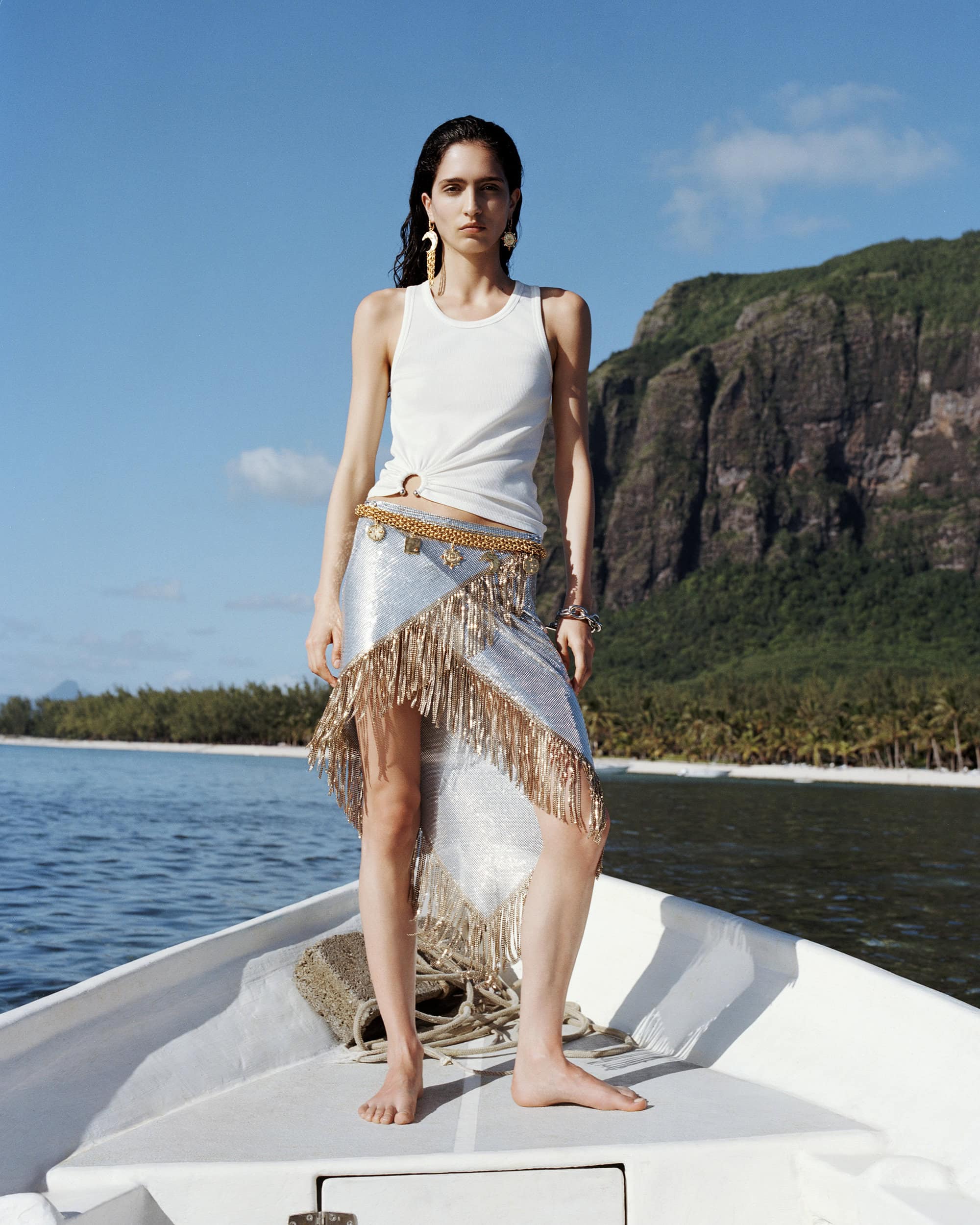 A woman in a white tank top and fringed skirt stands on a boat at sea with a mountain backdrop under a blue sky, creating a fashion editorial shot.