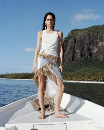 A woman in a white tank top and fringed skirt stands on a boat at sea with a mountain backdrop under a blue sky, creating a fashion editorial shot.