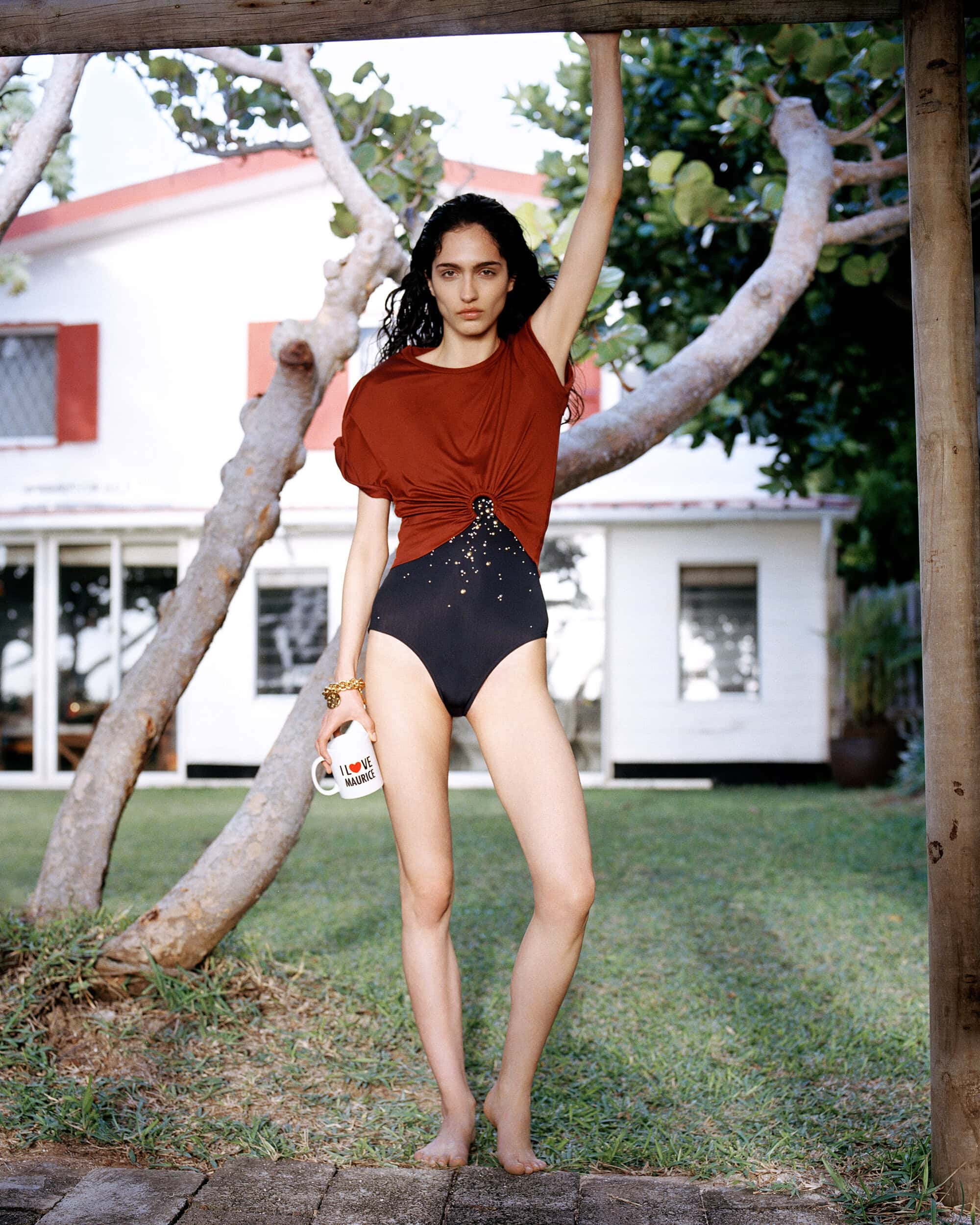 A woman in a swimsuit and top poses outside on a lawn with a white house and tree branches as a backdrop in a sunny, fashion-style portrait.