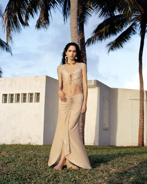 A young adult woman poses in a beige two-piece skirt and top set against a backdrop of palm trees and a white building on green grass.