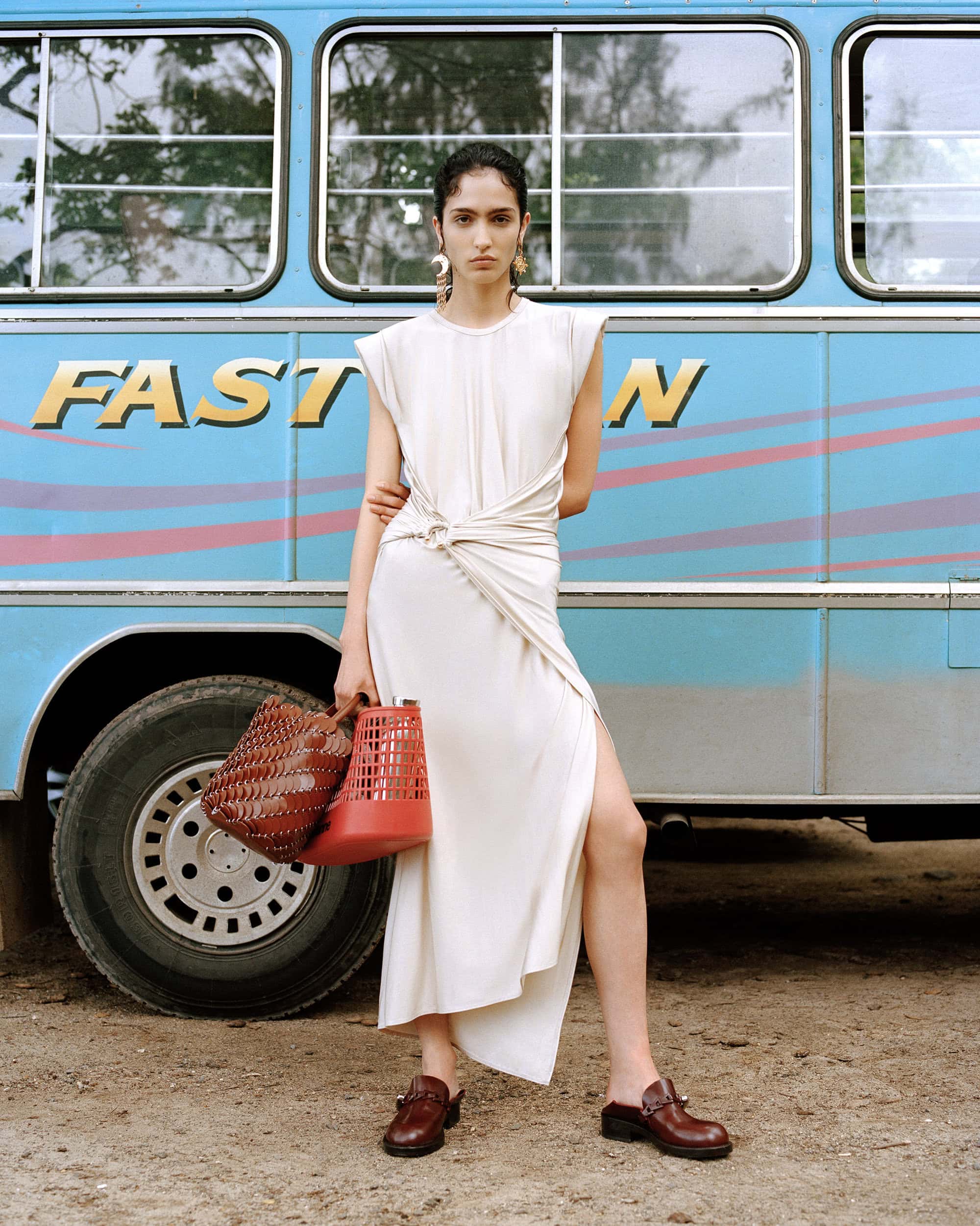 Model Sara Caballero wearing white dress holding woven bags in front of a blue bus for Rabanne Sundate campaign