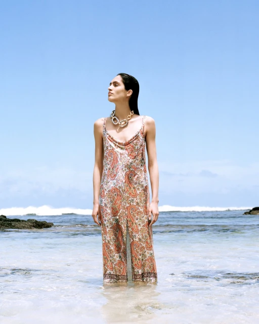 A woman in a paisley dress stands in shallow ocean water under a bright blue sky, accessorized with a statement gold necklace.