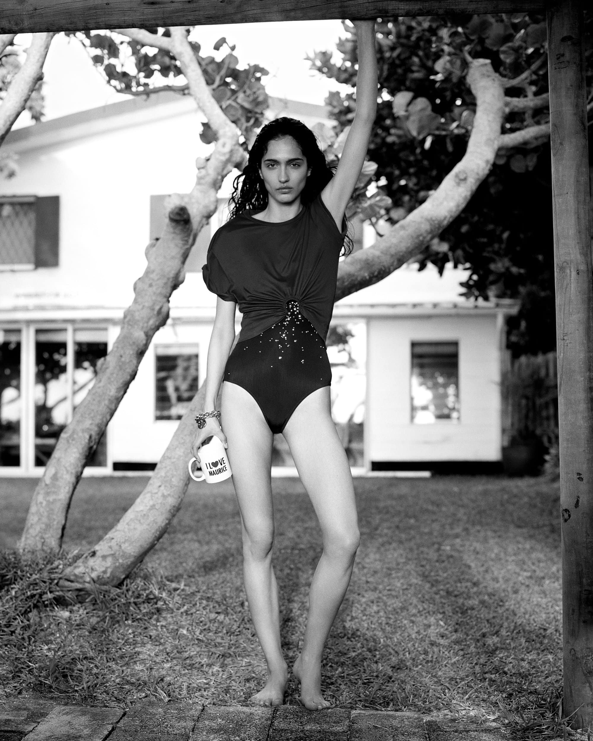 Black-and-white fashion shot of a slender woman in a swimsuit with a coffee cup, posing in front of a white house and trees.