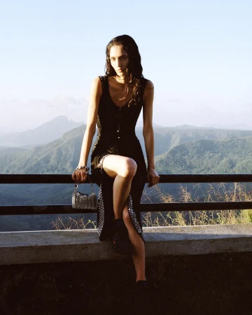 A woman in a black dress sits on a ledge with mountains in the background. She has a silver purse and is wearing dark boots.