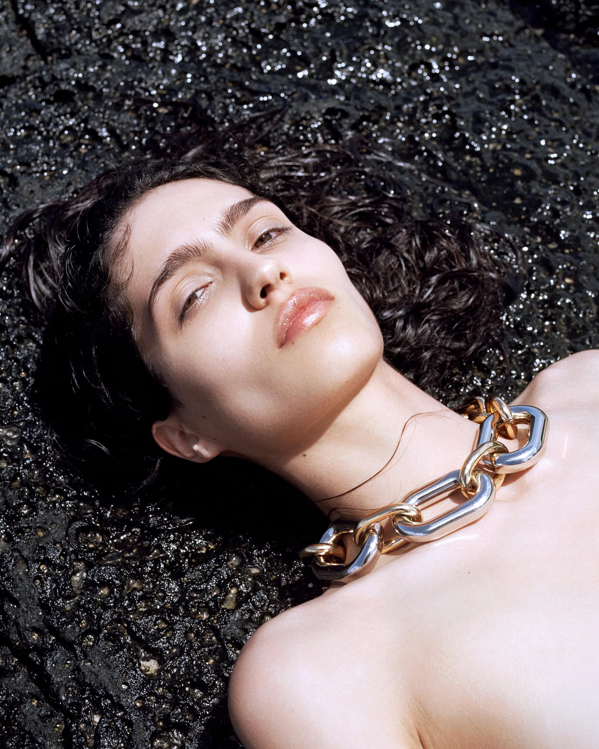 Close-up of an adult woman with curly dark hair and a silver and gold chain necklace, laying on a black rocky surface.