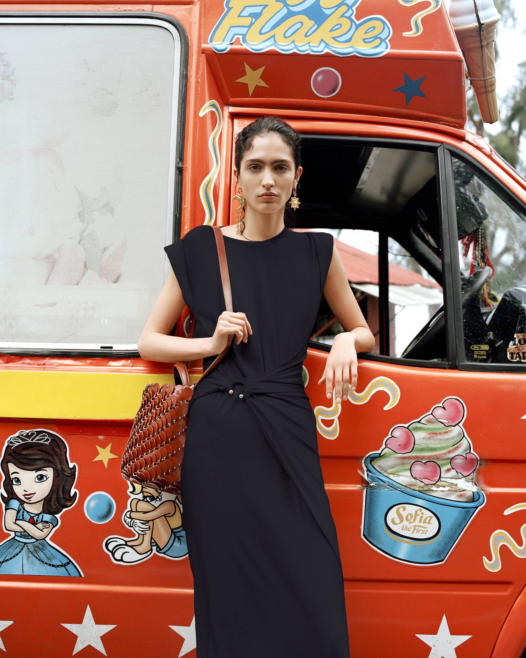 A slender woman in a black dress poses gracefully next to an eye-catching ice cream van, creating a striking and fashionable visual.