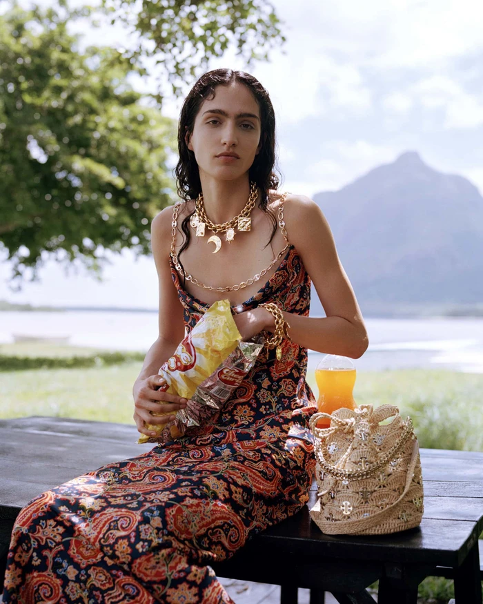 Model wearing a patterned dress and gold chain necklace, seated outdoors near water with a bucket bag and snacks.