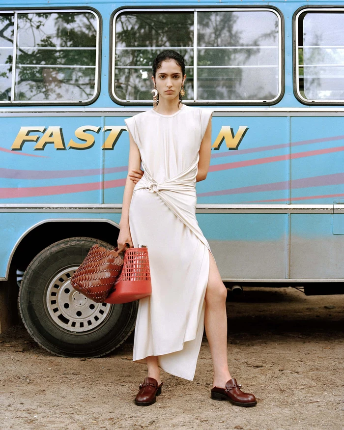 Model Sara Caballero wearing white dress holding woven bags in front of a blue bus for Rabanne Sundate campaign