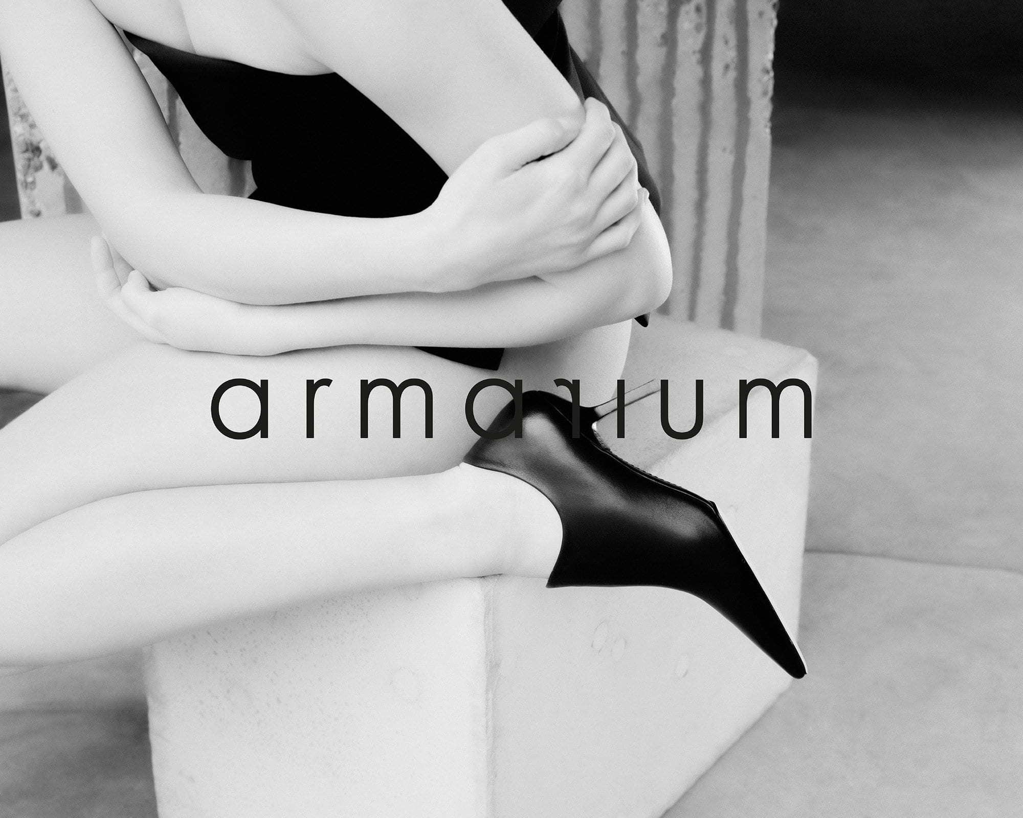 Woman wearing a black top and black heel sitting on a cubic stone block indoors, with an architectural column in the background.