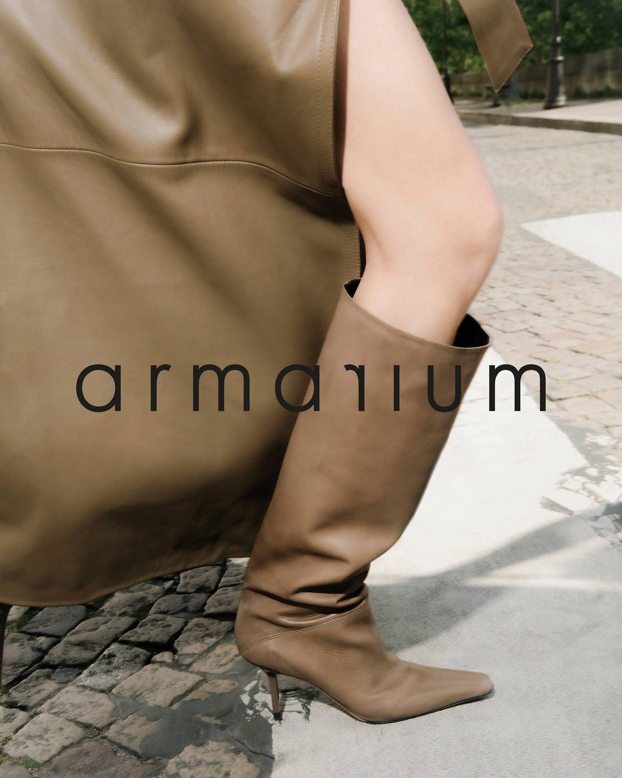 A woman's leg is shown wearing a knee-high, light brown leather boot, and the background includes cobblestones and a hint of greenery.